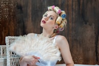 A woman with brightly colored curlers in her hair, wearing red lipstick and a pearl necklace, is posed in front of a textured, dark background. She is holding a large, fluffy feather fan that partially covers her chest. The wicker chair next to her adds to the vintage aesthetic.