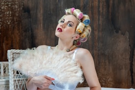 A woman with brightly colored curlers in her hair, wearing red lipstick and a pearl necklace, is posed in front of a textured, dark background. She is holding a large, fluffy feather fan that partially covers her chest. The wicker chair next to her adds to the vintage aesthetic.
