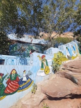 A vibrant mural on a wall features traditional Indian artwork. It includes colorful depictions of musicians playing instruments, dancers in cultural attire, and other folk elements. The backdrop includes detailed designs and additional smaller paintings. The mural is set amidst a natural environment with large trees and rock formations.