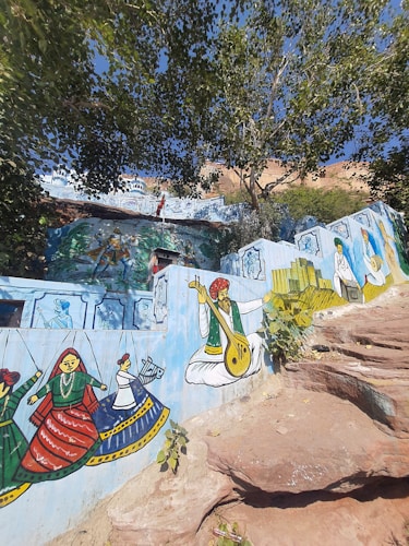 A vibrant mural on a wall features traditional Indian artwork. It includes colorful depictions of musicians playing instruments, dancers in cultural attire, and other folk elements. The backdrop includes detailed designs and additional smaller paintings. The mural is set amidst a natural environment with large trees and rock formations.