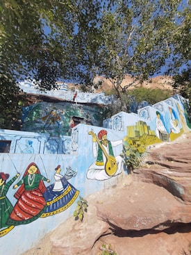 A vibrant mural on a wall features traditional Indian artwork. It includes colorful depictions of musicians playing instruments, dancers in cultural attire, and other folk elements. The backdrop includes detailed designs and additional smaller paintings. The mural is set amidst a natural environment with large trees and rock formations.