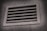 A rectangular metal vent with horizontal slats, embedded in a textured concrete wall, is shown, with a slight shadow cast around the vent frame.