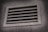 A rectangular metal vent with horizontal slats, embedded in a textured concrete wall, is shown, with a slight shadow cast around the vent frame.