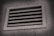 A rectangular metal vent with horizontal slats, embedded in a textured concrete wall, is shown, with a slight shadow cast around the vent frame.