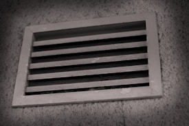 A rectangular metal vent with horizontal slats, embedded in a textured concrete wall, is shown, with a slight shadow cast around the vent frame.
