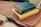 A stack of two books with leather covers labeled 'The Word of God' rests on a brown leather satchel. The books have blue and yellow covers, lying on a wooden surface. In the background, part of a white pot with a plant is visible.