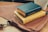 A stack of two books with leather covers labeled 'The Word of God' rests on a brown leather satchel. The books have blue and yellow covers, lying on a wooden surface. In the background, part of a white pot with a plant is visible.