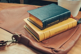 A stack of two books with leather covers labeled 'The Word of God' rests on a brown leather satchel. The books have blue and yellow covers, lying on a wooden surface. In the background, part of a white pot with a plant is visible.
