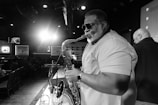 A musician with sunglasses plays the saxophone on stage in a dimly lit lounge. Other patrons are seen seated in the background against framed artwork. A spotlight creates a dramatic effect behind the performer.
