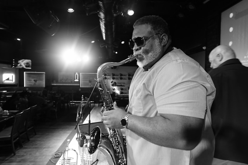 A musician with sunglasses plays the saxophone on stage in a dimly lit lounge. Other patrons are seen seated in the background against framed artwork. A spotlight creates a dramatic effect behind the performer.