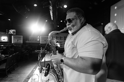 A musician with sunglasses plays the saxophone on stage in a dimly lit lounge. Other patrons are seen seated in the background against framed artwork. A spotlight creates a dramatic effect behind the performer.