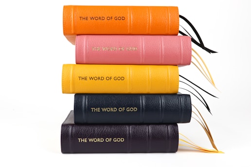 A stack of five books with colorful leather covers in orange, pink, yellow, dark blue, and black, embossed with the words 'THE WORD OF GOD' in gold lettering. They are placed on top of one another in a neat pile, with black and gold ribbon bookmarks visible.