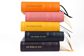 A stack of five books with colorful leather covers in orange, pink, yellow, dark blue, and black, embossed with the words 'THE WORD OF GOD' in gold lettering. They are placed on top of one another in a neat pile, with black and gold ribbon bookmarks visible.