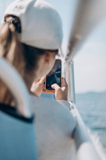 A person wearing a white cap is holding a smartphone and capturing a photo or video, with a blurred view of water in the background and an aluminum railing next to them.