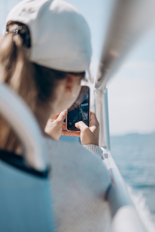 A person wearing a white cap is holding a smartphone and capturing a photo or video, with a blurred view of water in the background and an aluminum railing next to them.