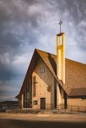 A modern church building with a tall, narrow steeple featuring a cross at the top. The structure is composed of light-colored bricks and large glass windows. The roof is steep and triangular, casting a shadow on the wall. The entryway is accessible by a ramp, and there are decorative elements like small crosses on the facade.