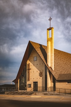 A modern church building with a tall, narrow steeple featuring a cross at the top. The structure is composed of light-colored bricks and large glass windows. The roof is steep and triangular, casting a shadow on the wall. The entryway is accessible by a ramp, and there are decorative elements like small crosses on the facade.