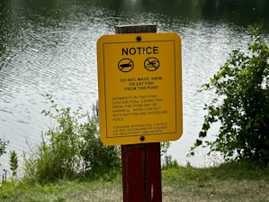 A yellow warning sign is displayed near a body of water, cautioning against swimming, wading, or eating fish due to the presence of harmful sediments. The background features rippling water and surrounding greenery, suggesting a natural, outdoor setting.