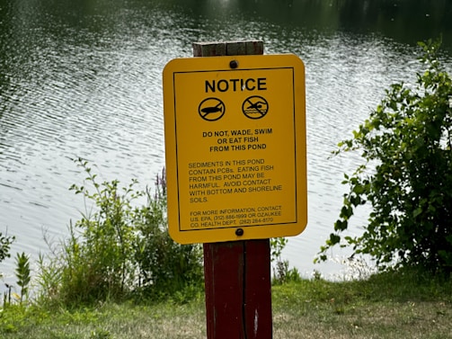 A yellow warning sign is displayed near a body of water, cautioning against swimming, wading, or eating fish due to the presence of harmful sediments. The background features rippling water and surrounding greenery, suggesting a natural, outdoor setting.