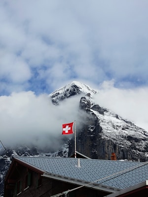 Snow-capped mountain peak emerging through clouds, with a Swiss flag prominently displayed on a pole in front of a wooden building topped with a blue roof.