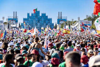 A large crowd of people is gathered outdoors, waving a variety of national flags. In the background, there is a modern building and two large screens displaying a speaker. The atmosphere appears festive and lively.