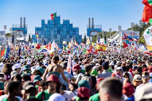 A large crowd of people is gathered outdoors, waving a variety of national flags. In the background, there is a modern building and two large screens displaying a speaker. The atmosphere appears festive and lively.