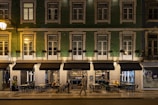 A street view of a building with green tiling and several windows illuminated by warm lighting. The ground floor houses a restaurant named Sebastião, with outdoor seating and patrons visible. One person is walking past the restaurant. The setting appears to be at night, as suggested by the artificial lighting and the ambience.