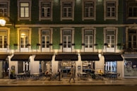 A street view of a building with green tiling and several windows illuminated by warm lighting. The ground floor houses a restaurant named Sebastião, with outdoor seating and patrons visible. One person is walking past the restaurant. The setting appears to be at night, as suggested by the artificial lighting and the ambience.