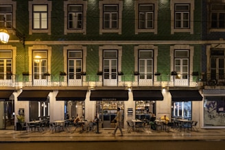 A street view of a building with green tiling and several windows illuminated by warm lighting. The ground floor houses a restaurant named Sebastião, with outdoor seating and patrons visible. One person is walking past the restaurant. The setting appears to be at night, as suggested by the artificial lighting and the ambience.