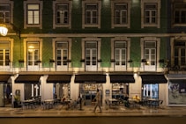 A street view of a building with green tiling and several windows illuminated by warm lighting. The ground floor houses a restaurant named Sebasti&atilde;o, with outdoor seating and patrons visible. One person is walking past the restaurant. The setting appears to be at night, as suggested by the artificial lighting and the ambience.