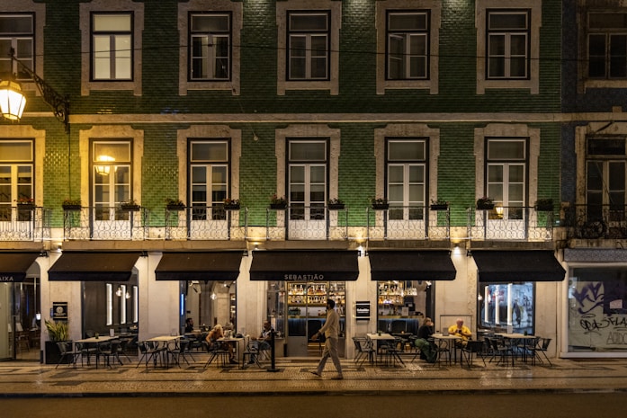A street view of a building with green tiling and several windows illuminated by warm lighting. The ground floor houses a restaurant named Sebastião, with outdoor seating and patrons visible. One person is walking past the restaurant. The setting appears to be at night, as suggested by the artificial lighting and the ambience.