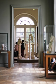 A museum gallery with classical sculptures displayed on pedestals near a large arched window. Two people are observing the art, and there are additional sculptures in glass cases on either side of the room.