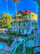 A multi-story building with a red roof and green shutters surrounded by lush vegetation. There is a balcony with green railings in the foreground and a swimming pool below. Palm trees and other greenery fill the landscape, while bright daylight highlights a vibrant blue sky.