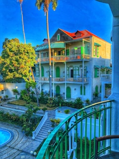 A multi-story building with a red roof and green shutters surrounded by lush vegetation. There is a balcony with green railings in the foreground and a swimming pool below. Palm trees and other greenery fill the landscape, while bright daylight highlights a vibrant blue sky.