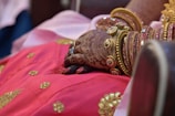 A hand adorned with elaborate henna designs is seen wearing several gold and pink bangles, resting on a vibrant pink garment with intricate gold embroidery.