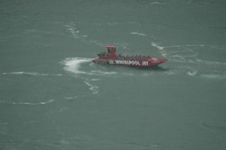 A red jet boat named 'Whirlpool Jet' speeds through a body of water, creating a white wake behind it. The boat is filled with people wearing blue life jackets.