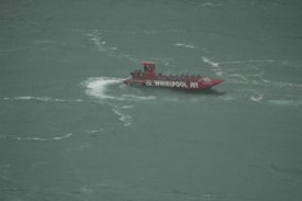 A red jet boat named 'Whirlpool Jet' speeds through a body of water, creating a white wake behind it. The boat is filled with people wearing blue life jackets.