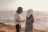 Two people stand facing each other near a beach with a large body of water and a sunset in the background. Both are wearing Star Wars helmets; one is dressed in a white short-sleeved shirt and black pants, and the other is in a denim jacket.