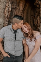 A couple is standing close together against a large tree trunk. The man is wearing a gray short-sleeve shirt and jeans, leaning slightly to kiss the woman on her forehead. She has long, wavy reddish-brown hair and is wearing an off-shoulder light pink dress with small floral patterns. They both appear happy and content.