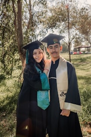 Two individuals wearing graduation caps and gowns are standing close together, smiling at the camera. They are outdoors in a park-like setting with trees and greenery surrounding them. The sunlight is filtering through the trees, creating a warm and cheerful atmosphere.