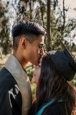 A young person gently kisses the forehead of another individual who is wearing a graduation cap and gown. They are outdoors in a lush, green environment with trees in the background, and the scene is bathed in warm sunlight.