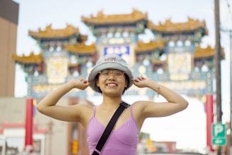 A young person wearing glasses, a bucket hat, and a purple top stands smiling with arms raised in front of a traditional Asian-style archway. The archway features ornate decorations and vibrant colors. The background includes elements of a busy street with various signs and buildings.