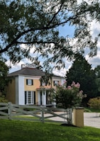 A two-story house with a beige exterior and white columns at the entrance. The house is surrounded by a neatly maintained lawn and various trees, including some with pink blossoms. There is a white wooden fence in the foreground and a driveway leading up to the house. The sky is partly cloudy with patches of blue visible.