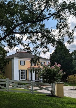 A two-story house with a beige exterior and white columns at the entrance. The house is surrounded by a neatly maintained lawn and various trees, including some with pink blossoms. There is a white wooden fence in the foreground and a driveway leading up to the house. The sky is partly cloudy with patches of blue visible.