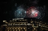 Bright fireworks illuminate the night sky above a grand, well-lit building identified as The Fullerton Hotel. The scene is vibrant with red and blue explosions contrasting against the dark background.