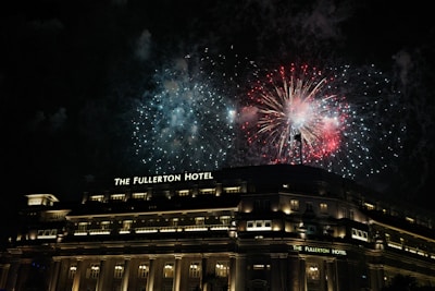 Bright fireworks illuminate the night sky above a grand, well-lit building identified as The Fullerton Hotel. The scene is vibrant with red and blue explosions contrasting against the dark background.