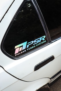 A close-up view of a car window displaying a decal with the text 'PULSAR TURBO SYSTEMS' and the logo 'PSR' in a gradient color that shifts from pink to blue. The window is part of a white vehicle.