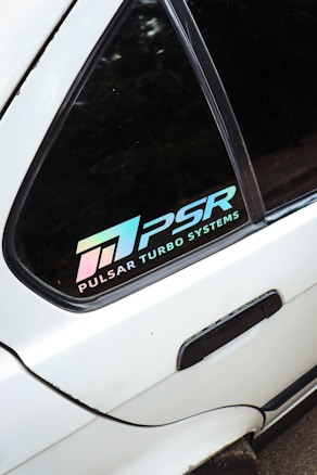 A close-up view of a car window displaying a decal with the text 'PULSAR TURBO SYSTEMS' and the logo 'PSR' in a gradient color that shifts from pink to blue. The window is part of a white vehicle.