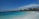 A picturesque beach scene featuring turquoise waters gently lapping against the white sandy shore. Thatched-roof huts and palm trees line the background under a blue sky with scattered clouds. People can be seen enjoying the water and relaxing along the shore.