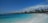 A picturesque beach scene featuring turquoise waters gently lapping against the white sandy shore. Thatched-roof huts and palm trees line the background under a blue sky with scattered clouds. People can be seen enjoying the water and relaxing along the shore.